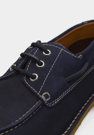 Chaussure bateau en daim bleu marine avec coutures blanches, lacets noirs passés dans des œillets métalliques, et une doublure intérieure en cuir marron.