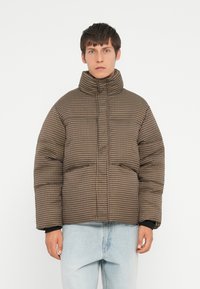 Bruin en beige geruite pufferjack met een hoge kraag, knopen aan de voorkant en twee zijzakken, gecombineerd met lichtblauwe jeans.