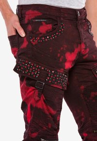 Rote und schwarze, stark abgenutzte Denim-Jeans mit metallischen Nieten, Nieten und einer strukturierten Oberfläche. Verfügt über dekorative Beschläge und einzigartige Muster.