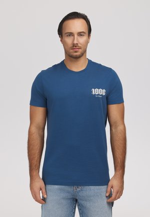 Mężczyzna z krótkimi ciemnymi włosami, ubrany w niebieską koszulkę "1908 Lee Cooper" i jasnoniebieskie dżinsy, stojący na jednolitym tle.