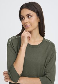 Femme aux longs cheveux noirs portant un pull vert olive avec des détails de boutons sur les épaules, regardant sur le côté avec une main près du menton.