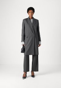 Cappotto grigio a righe con un grande colletto, abbinato a pantaloni a gamba larga. Presenta maniche lunghe con dettagli a coste e una piccola borsa nera.