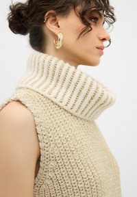 Maglione beige lavorato a maglia con un collo a dolcevita spesso e rigato. La trama è corposa, con riflessi di luce che mettono in risalto l'intreccio dettagliato. Orecchini a cerchio dorati.