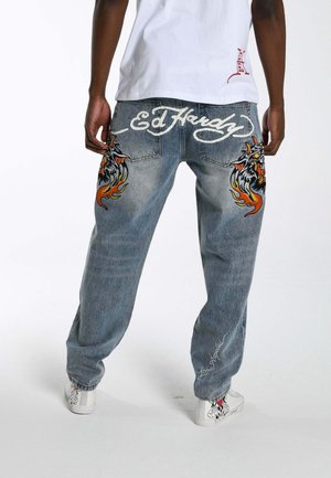 Person trägt blaue Jeans mit feurigen Tiermotiven auf den Oberschenkeln, "Ed Hardy"-Logo auf der Rückseite und "Los Angeles"-Schriftzug in der Nähe des Knöchels.