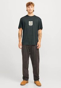 Camiseta de algodón negra de manga corta, con un gráfico central en color crema. Combinada con jeans de mezclilla marrones oscuros y zapatos color beige.