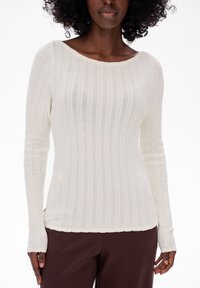 Sweter z długim rękawem w kolorze kremowym, z szerokim dekoltem w łódź i skomplikowanym wzorem teksturalnym, w połączeniu z luźnymi brązowymi spodniami.