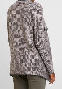 Lehký cardigan v herringbone vzoru v šedých a béžových odstínech, s křivkovým lemem a volným střihem, doplněný viditelnými švy.