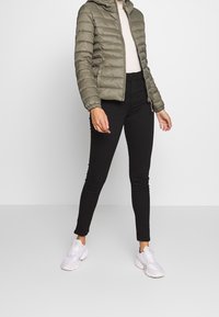 Oliwkowa puchowa kurtka z kapturem o pikowanej teksturze, z suwakiem z przodu i elastycznymi mankietami, zestawiona z czarnymi rurkami i białymi trampkami.