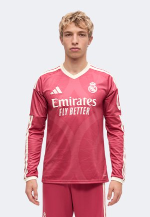 Giovane uomo con maglia da calcio a maniche lunghe rossa del Real Madrid con il logo Emirates Fly Better, in piedi davanti a uno sfondo chiaro uniforme.