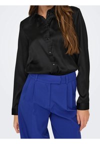 Chemise noire en satin à manches longues avec un col à boutons, associée à un pantalon plissé bleu vif taille haute. Texture lisse, coupe ajustée.