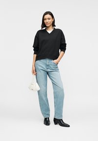 Μαύρο φούτερ με λαιμόκοψη V, ανοιχτό μπλε τζιν με ίσια γραμμή, μαύρα μποτάκια με τακούνι και λευκή τσάντα χειρός με υφή. Μοντέλο σε ουδέτερο φόντο.