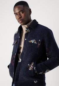 Ναυτικό μπλε μπουφάν με υφαντή επιφάνεια, με πολύχρωμα σχέδια, αποσπώμενο κολάρο από σέρπα και μπροστινές τσέπες. Τα χαρακτηριστικά περιλαμβάνουν κουμπιά και φερμουάρ.