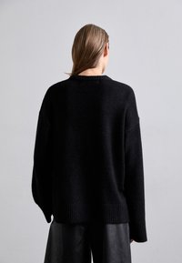 Musta neulepusero, jossa väljä istuvuus ja teksturoitu materiaali. Varustettu resorireunuksilla helmassa ja hihansuut, pyöreällä pääntiellä ja lasketuilla harteilla.