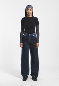 Zwarte aansluitende t-shirt over een lange mouwen grijze top, gecombineerd met wijde donkerblauwe jeans en zwarte instapschoenen.