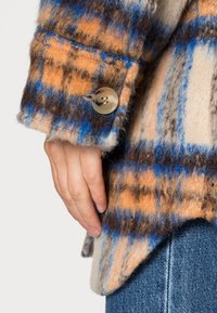 Rutig ulljacka med orange, blå och bruna mönster. Anmärkningsvärda detaljer inkluderar en framträdande knapp och en mjuk, texturerad yta.