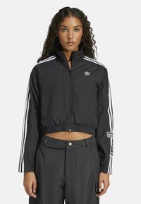 Jachetă neagră din material windbreaker, cu accente albe formate din trei dungi. Dispune de un fermoar complet, guler în picioare și un logo brodat pe piept.