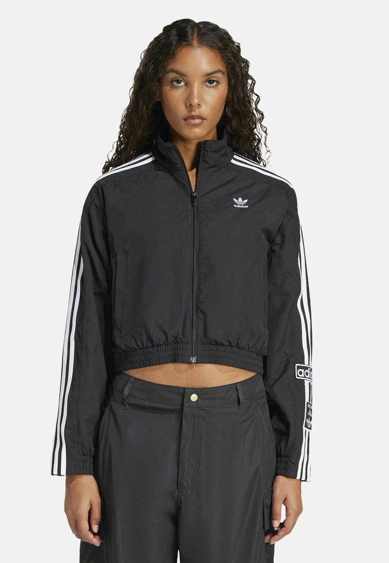 Jachetă neagră din material windbreaker, cu accente albe formate din trei dungi. Dispune de un fermoar complet, guler în picioare și un logo brodat pe piept.