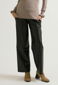 Schwarze Kunstleder-Breit-Bein-Hosen mit frontalen Falten, kombiniert mit einem beigefarbenen, gerippten Pullover mit Rüschenmanschetten und Knopfakzenten.