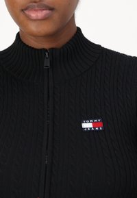 Melnā ribota ar rāvējslēdzēju aizdares džemperis ar augstu apkakli. Iezīmēts ar kabeli adījuma dizainu un mazu "Tommy Jeans" emblēmu sarkanā, baltā un zilā krāsā.