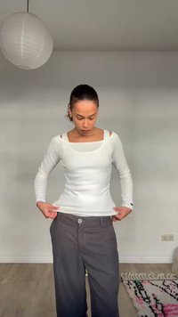 Ženska z zglajenimi lasmi nosi belo majico z dolgimi rokavi in temne široke hlače ter si popravlja srajco v minimalistični sobi z visečo svetilko.