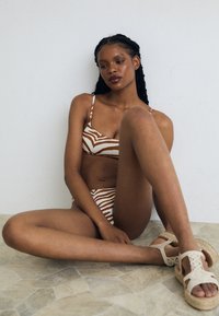 Bikini komplet z zebrastripes v rjavih in bež odtenkih, s tankimi naramnicami. V kombinaciji z pletenimi bež sandalami na teksturiranem tleh.