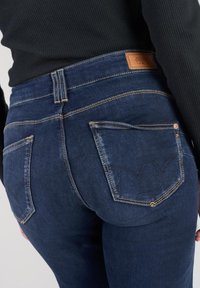 Los vaqueros de mezclilla azul oscuro tienen una cintura alta, un sutil desgaste y dos bolsillos traseros con un diseño de costura distintivo. Parche de marca de cuero visible.