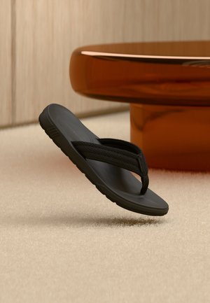Schwarze Flip-Flop-Sandale mit strukturiertem Riemen, die über einem beigen Teppich neben einem runden, bernsteinfarbenen Tischfuß schwebt.