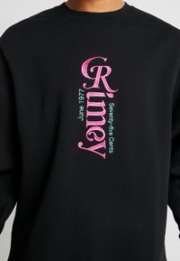 Felpa nera con ricamo verticale che riporta la scritta "Rimey" in rosa, e "Giugno 1977" e "Settantacinque centesimi" in azzurro chiaro.