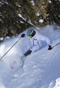 Skieur en combinaison isolante blanche avec des accents noirs, casque vert et bâtons de ski, taillant à travers la neige profonde dans une zone boisée.