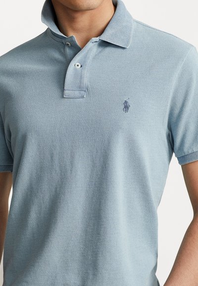 Gaiši zils polo krekls, kas izgatavots no teksturētas auduma. Tam ir klasiskā apkakle, divu pogu aizdare un izšūts logotips uz krūšu daļas.