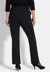 Vrouw in zwarte rechte broek en zwarte open teen hoge hakken, staand met haar rug naar de kijker toe tegen een effen achtergrond.