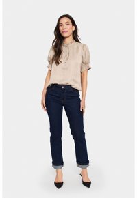 Blusa di satin beige con maniche a sbuffo e colletto a volant, abbinata a jeans blu scuro con risvolti e tacchi a punta neri.