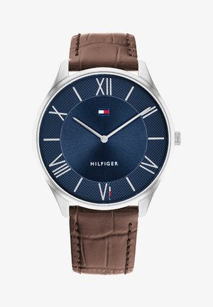Reloj plateado con una esfera texturizada en azul marino, marcadores de números romanos y correa de piel de cocodrilo marrón. Logotipo "HILFIGER" mostrado.