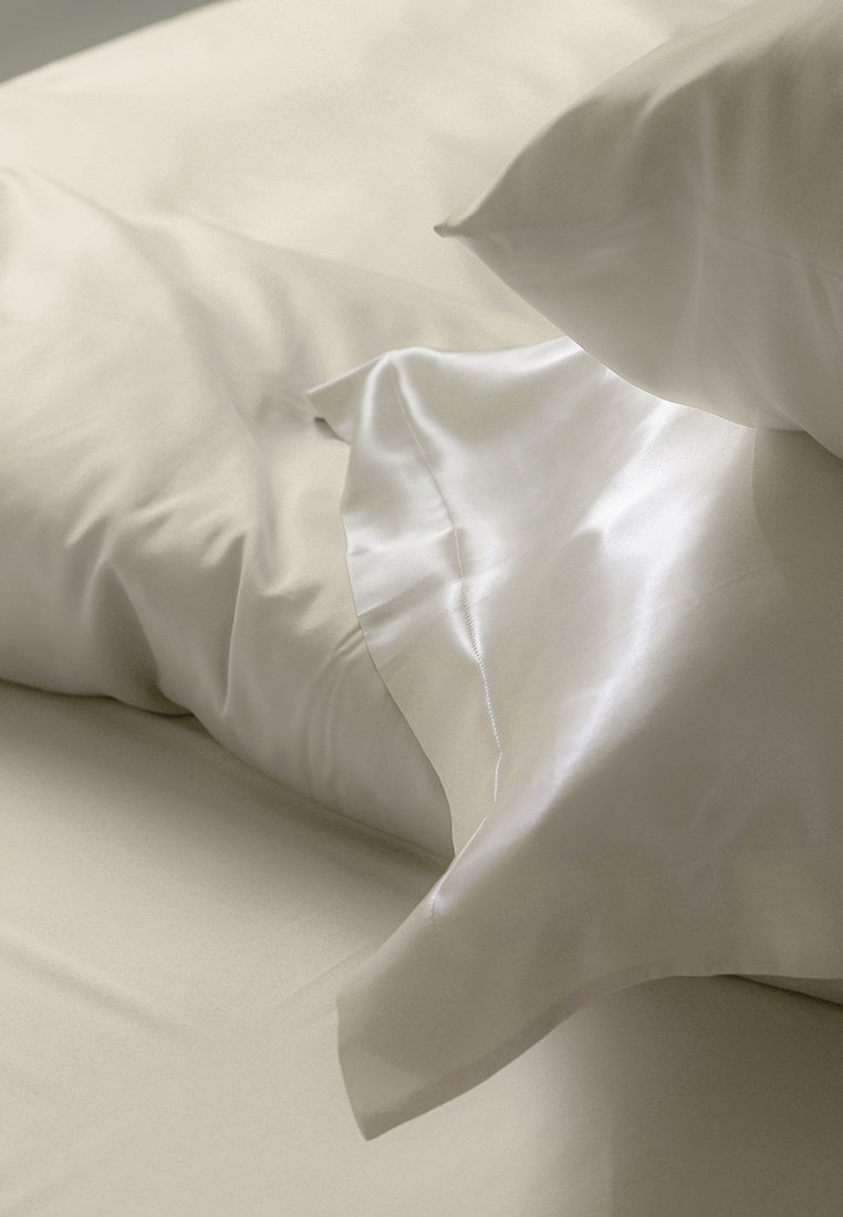 Weiße Baumwoll- und Satin-Kissenbezüge, die auf beigen Bettlaken lagen, mit glatten Texturen und einem einfachen, eleganten Design mit klaren Linien.