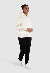 Cremefarbener Kapuzenpullover mit Kängurutasche, kombiniert mit schwarzen Jogginghosen und weißen Turnschuhen. Glatter Stoff, lockere Passform, minimales Branding.