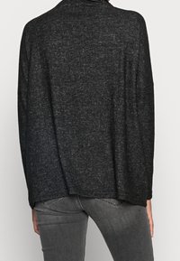 Maglione nero, textured con un fit morbido, maniche larghe e un leggero orlo asimmetrico, indossato sopra jeans grigi.
