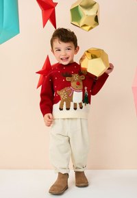 Pull en tricot rouge avec motif de renne et bas blanc, associé à un pantalon en velours côtelé crème et des bottes beiges, sur fond de décorations géométriques colorées.