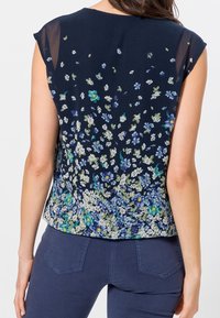 Šaty bez rukávů z námořnické tkaniny s květinovým vzorem, který má modré, zelené a bílé květy, a s průsvitným přehozem na ramenou.