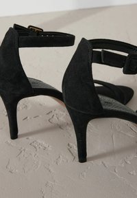 Sandalias de tacón alto de ante negro con punta afilada, correa para el tobillo y suela texturizada. Presentan un tacón de aguja y un diseño minimalista.