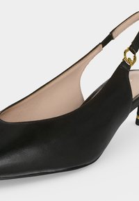 Chaussure slingback en cuir noir avec petit talon kitten, doublure intérieure beige et détail boucle métallique dorée sur la bride.