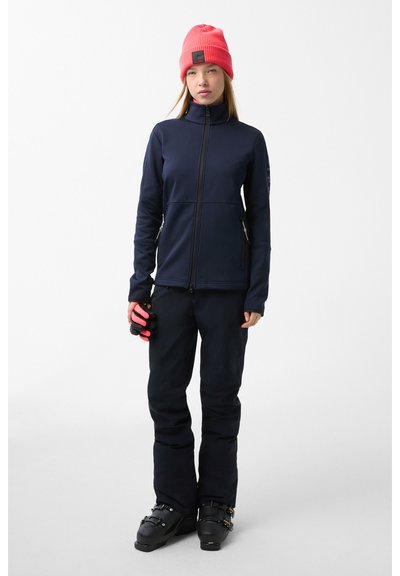 Navy softshell jacket with a high collar, zippered pockets, and pink beanie. Black ski pants and boots complete the outfit.