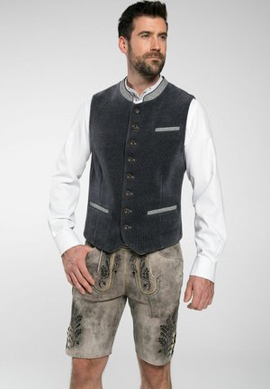 Graue Cordweste mit Frontknöpfen, kombiniert mit beigen Shorts, die über einen floralen Stickerei und dekorative Nähte verfügen. Weißes Hemd darunter.