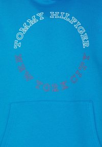 Sudadera azul con un diseño circular que presenta "TOMMY HILFIGER" en blanco y "NEW YORK CITY" en rojo, con un bolsillo frontal.