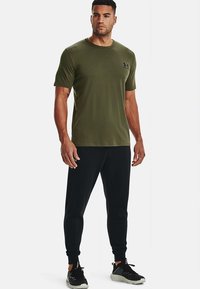 Mand stående iført en olivengrøn T-shirt, sorte løbebukser og sorte sneakers med hvide såler på en hvid baggrund.