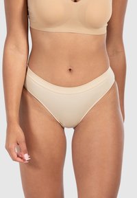 Donna che indossa biancheria intima alta a costine color beige e un reggiseno senza cuciture abbinato, in piedi contro uno sfondo grigio chiaro uniforme.