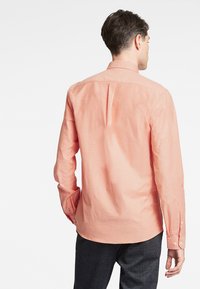Lange mouw knoopshirt in licht perzik katoen met een rugplooi en knoopmanchetten, gecombineerd met donkere gestructureerde broek.