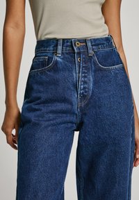 Jeans de ganga azul de cintura alta com um corte relaxado, apresentando costuras em contraste, bolsos frontais e fecho de botão único.