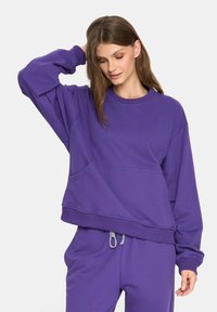 Kobieta w oversize'owej fioletowej bluzie i dopasowanych fioletowych dresach ze sznurkiem, pozująca z jedną ręką na głowie i patrząca w dół.