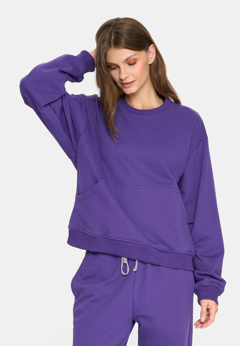 Kobieta w oversize'owej fioletowej bluzie i dopasowanych fioletowych dresach ze sznurkiem, pozująca z jedną ręką na głowie i patrząca w dół.