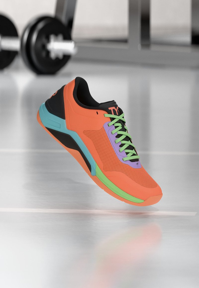 Φωτεινό πορτοκαλί αθλητικό παπούτσι με πράσινα κορδόνια, μοβ και πράσινες λεπτομέρειες, τοποθετημένο σε γυαλιστερό πάτωμα γυμναστηρίου με βάρη στο φόντο.